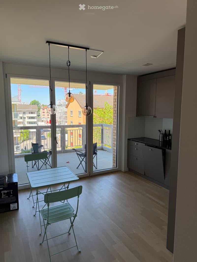 Modern kitchen with wooden flooring, grey cabinets, and a small dining area with a table and chairs. A sliding glass door opens to a balcony with additional seating.