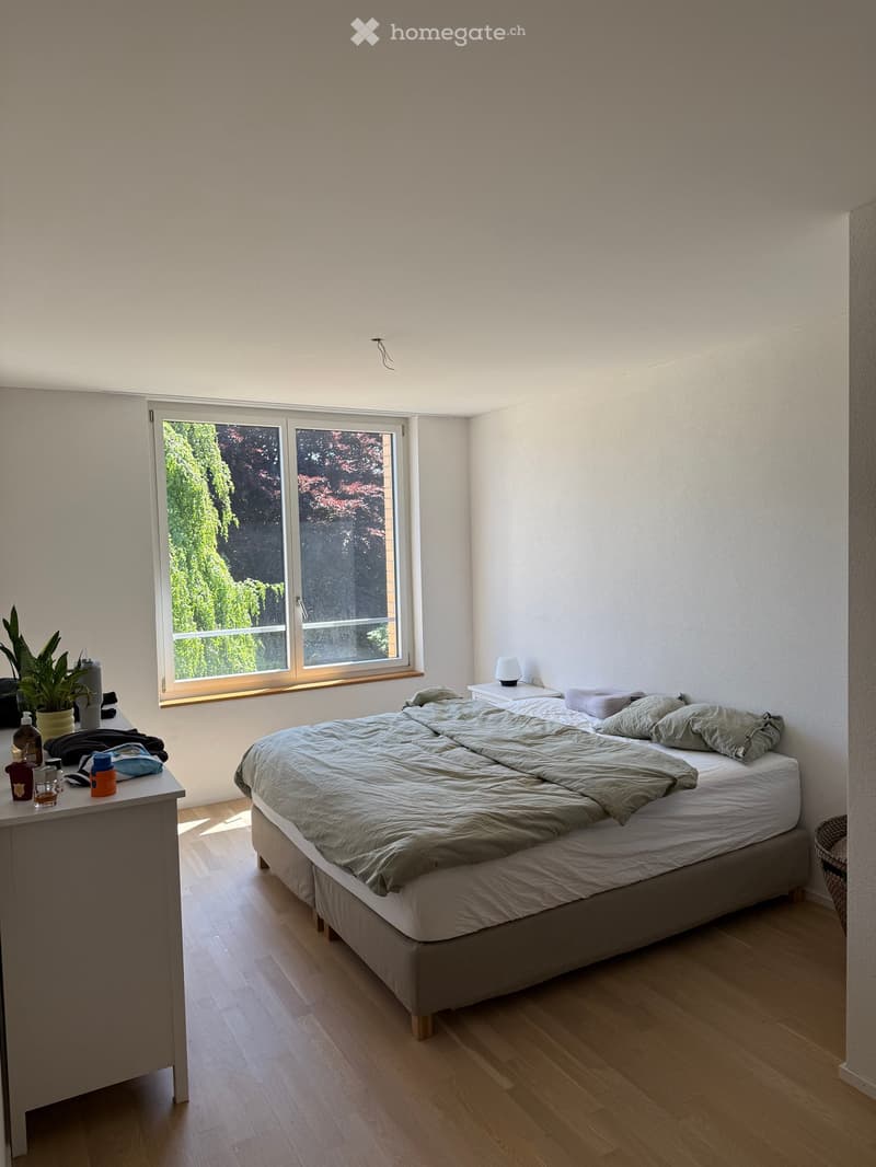 A modern, minimalist bedroom with a large bed, nightstand, lamp, and a small table with a plant and various items.