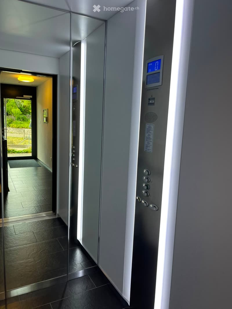 Modern hallway with dark tiled flooring, a sleek elevator with digital display and control panel, and a view of a bright, open area beyond a glass door.