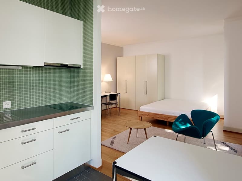 Modern studio apartment with a kitchen area featuring white cabinets and an induction stove, a bedroom area with a bed and wardrobe, a desk with a chair, and a small seating area with a stool and a teal chair.