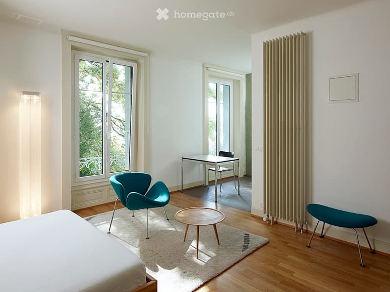 Bright, minimalist room with a bed, teal chairs, a small table, and a radiator. Large windows allow natural light to fill the space.