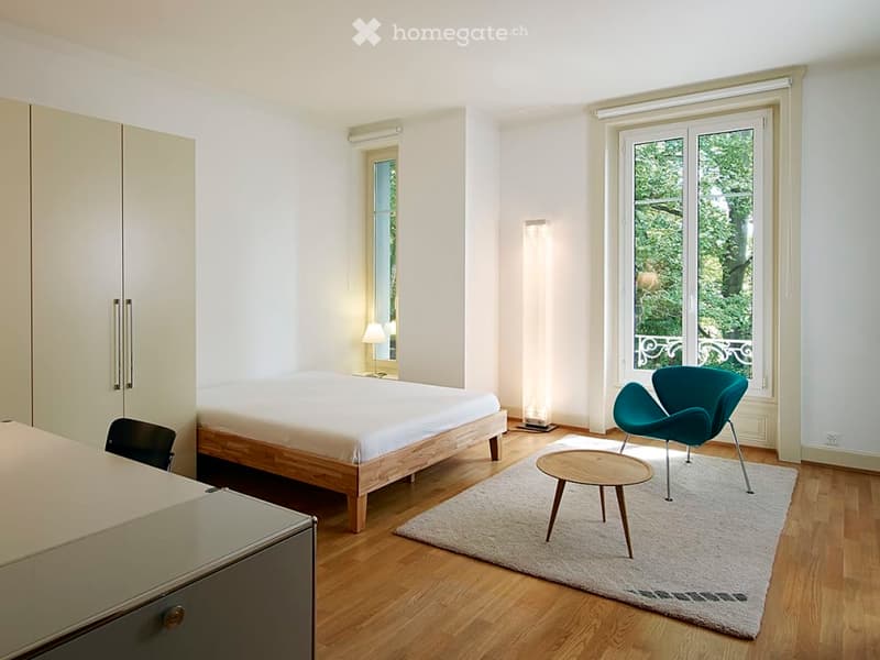 Modern bedroom with wooden flooring, a bed, a teal chair, a small table, a wardrobe, and a lamp near the window.