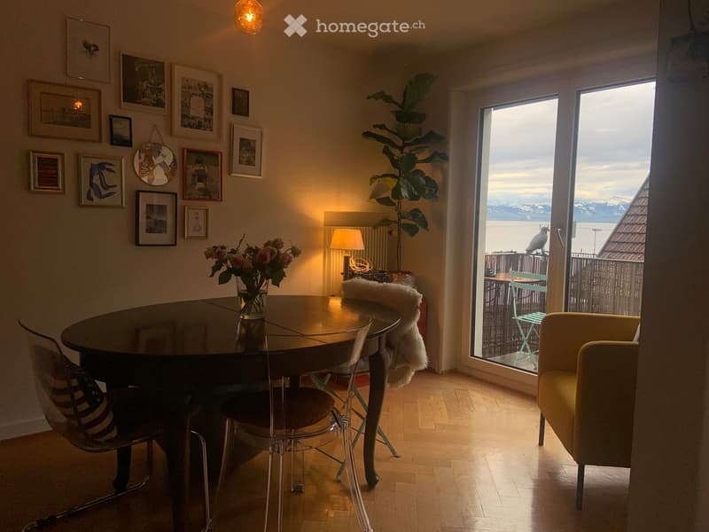 Bright dining area with a round wooden table, chairs, a lamp, and a vase with flowers. Large windows open to a balcony with outdoor seating.