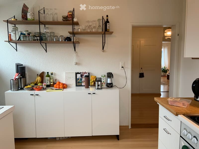 Modern kitchen with white cabinets, wooden countertops, shelves with glassware and jars, coffee maker, and various kitchen appliances.
