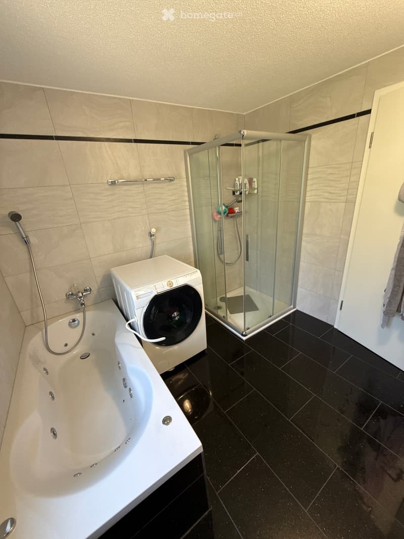 Modern bathroom with a bathtub, glass-enclosed shower, and a private washing machine. Features include a towel rack and a towel on the side.