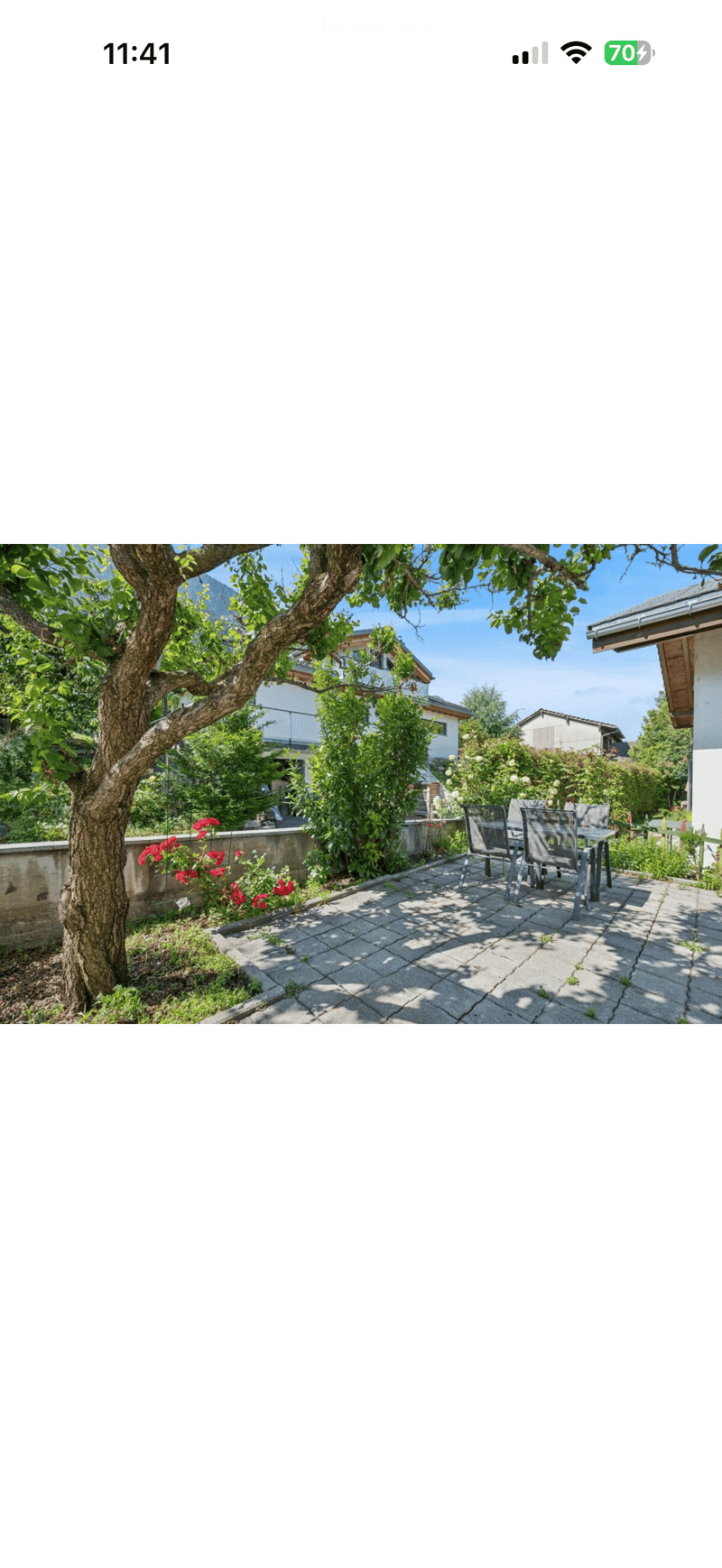 A sunny outdoor patio with a table and chairs, surrounded by lush greenery and flowers, with a large tree providing shade.