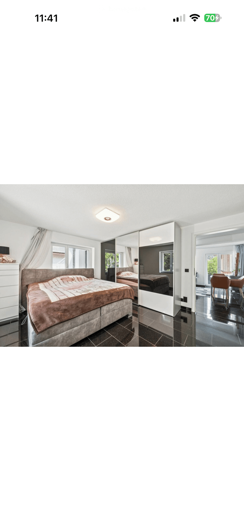 Modern bedroom with a large bed, mirrored wardrobe, and a view into an adjacent living area with a dining table and chairs.