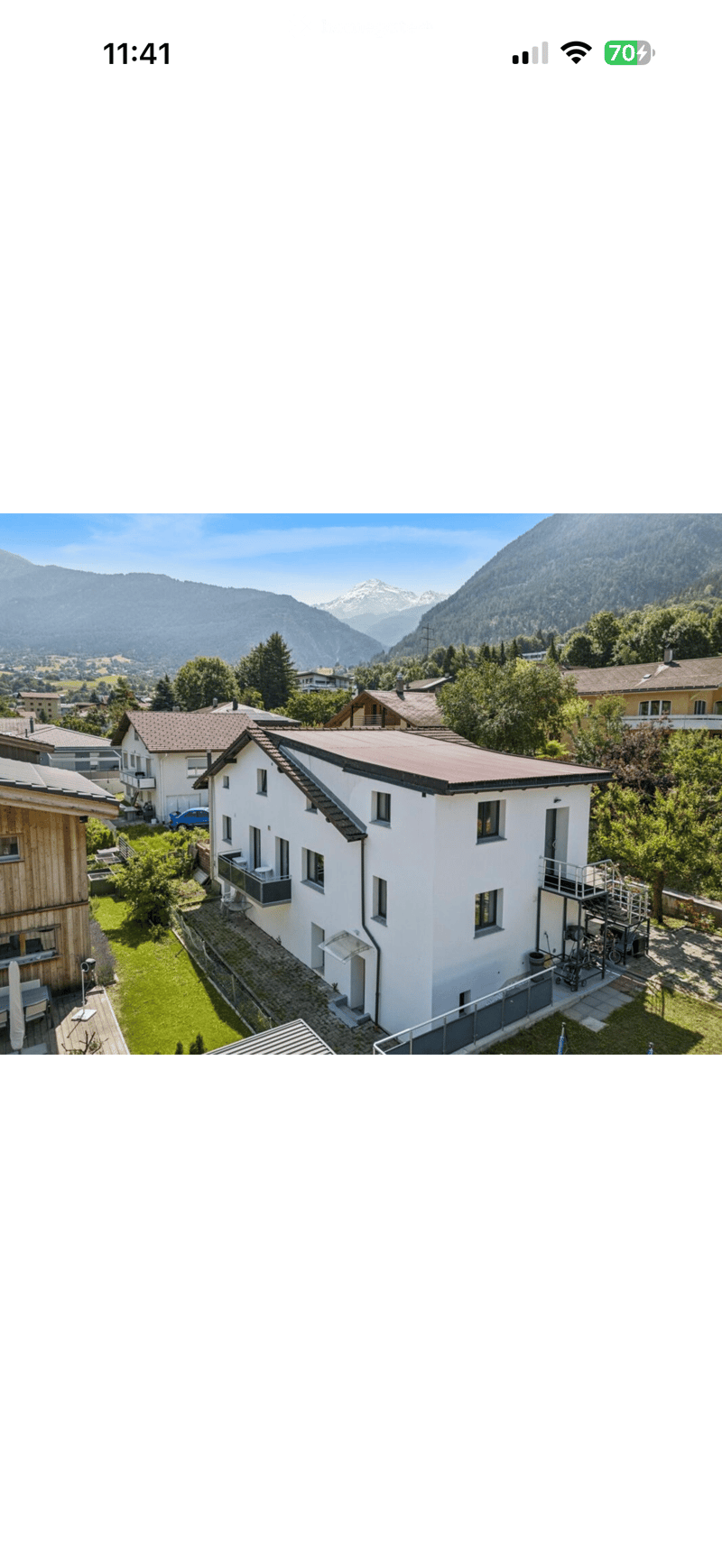 A modern two-story house with a red-tiled roof, white exterior, and large windows, situated in a scenic mountainous area with neighboring houses and lush greenery.