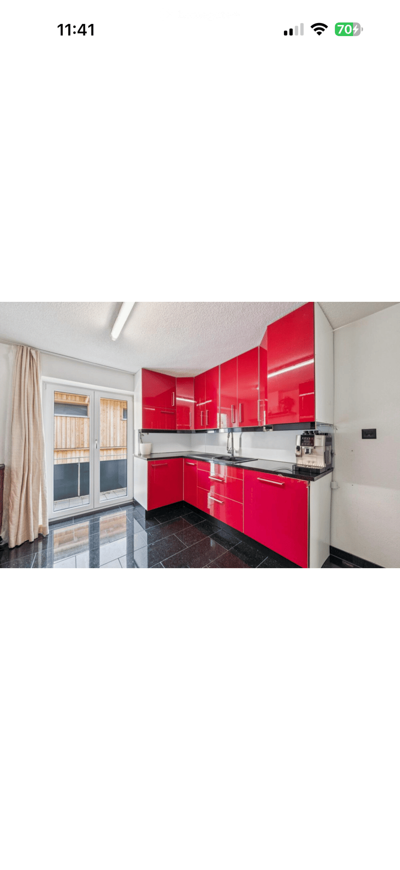 Modern kitchen with red cabinets, black countertops, and a stainless steel sink. Features a coffee machine and a window with a curtain.