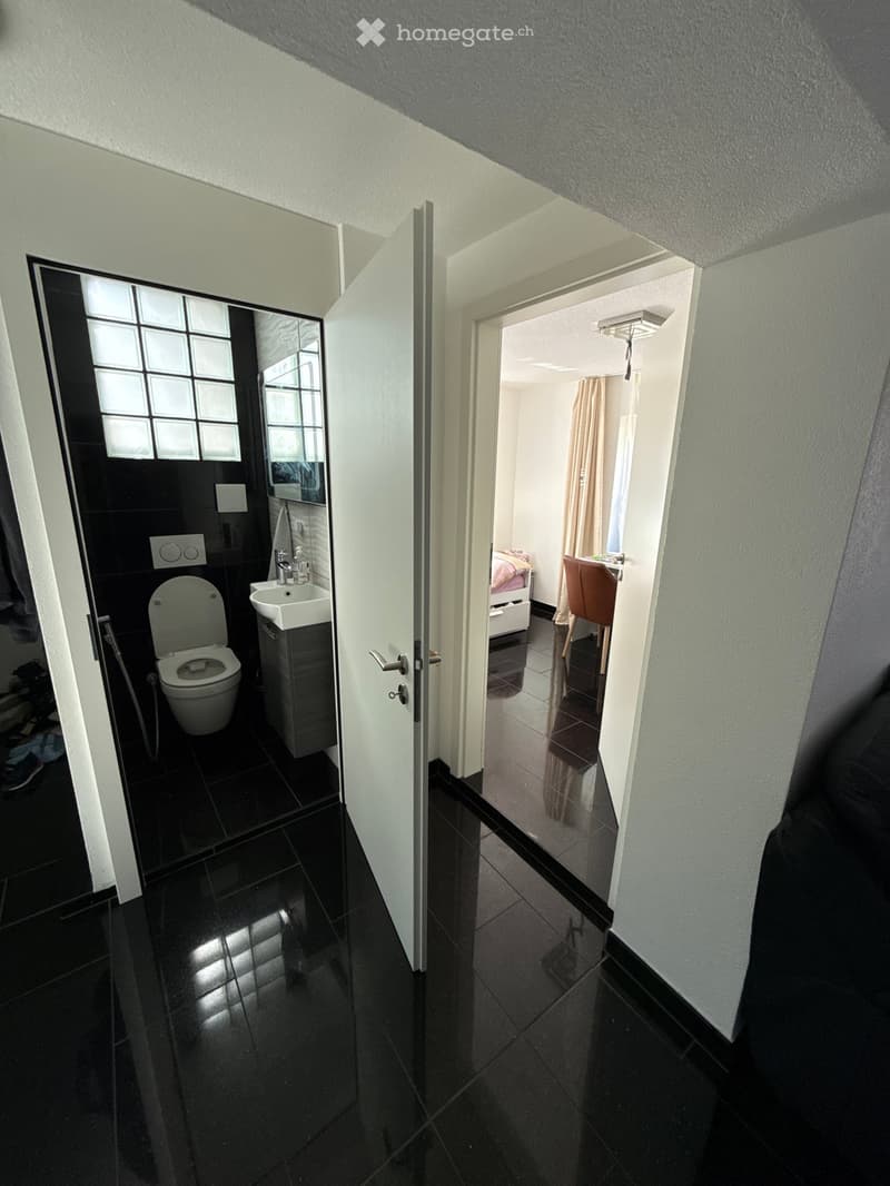 A narrow hallway with black tiled flooring and white walls, leading to a small bathroom and a bedroom. The bathroom features a toilet, sink, and mirror, while the bedroom has a bed, chair, and curtain.