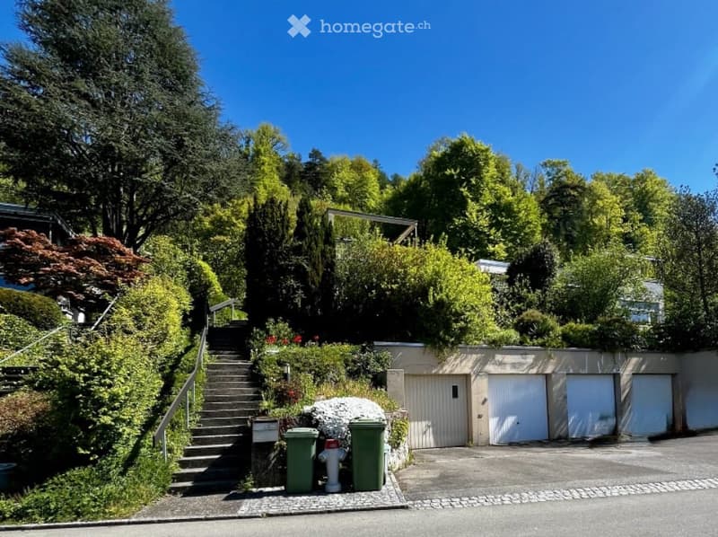 A house with a garage and a driveway, surrounded by lush greenery and trees.