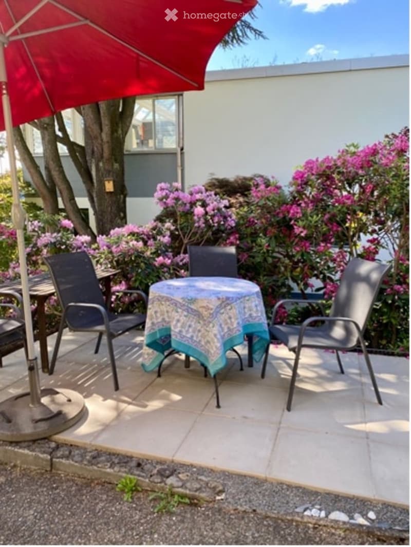 Outdoor seating area with a table and chairs, a red umbrella, and a tablecloth. Surrounded by pink flowers and a tree.