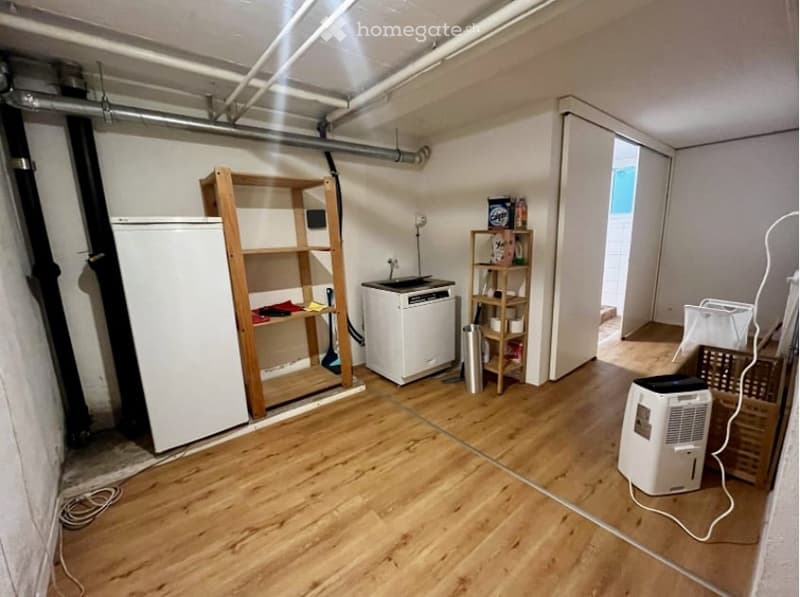 An empty room with wooden flooring and white walls. There are two wooden shelves, a white refrigerator, a white washing machine, a wooden storage box, and a white air conditioner. There are also some pipes and wires on the ceiling.
