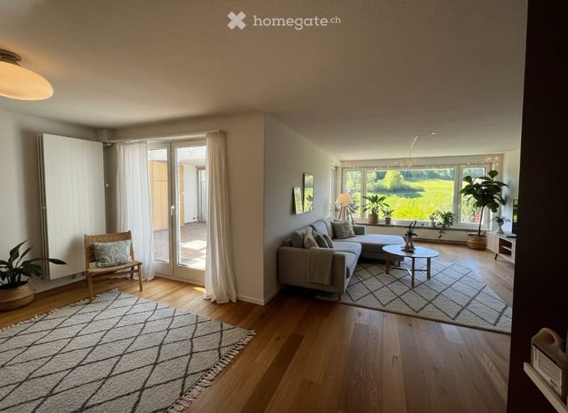 Spacious living room with wooden floors, large windows, and sliding glass doors leading to a balcony. The room features a gray couch, a coffee table, and a rug. There are potted plants and a lamp on the side table. The room is well-lit with natural light