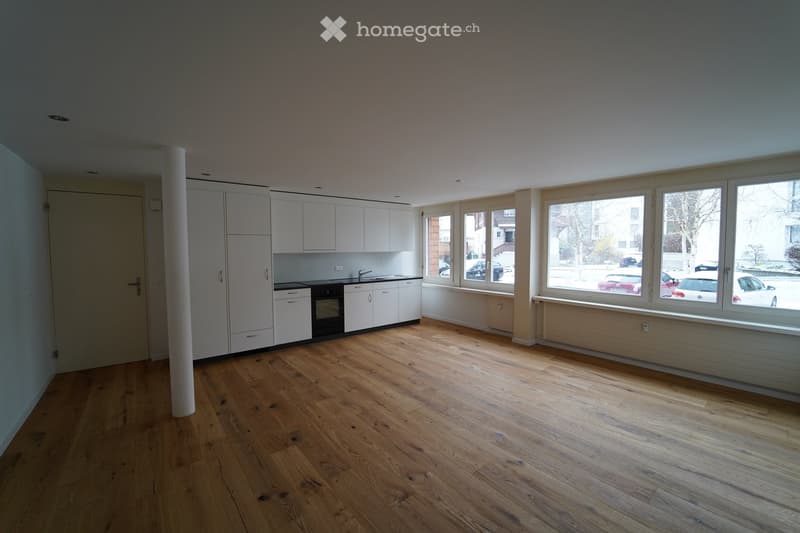 Empty room with light wooden flooring, white cabinetry, and large windows overlooking a street with parked cars.