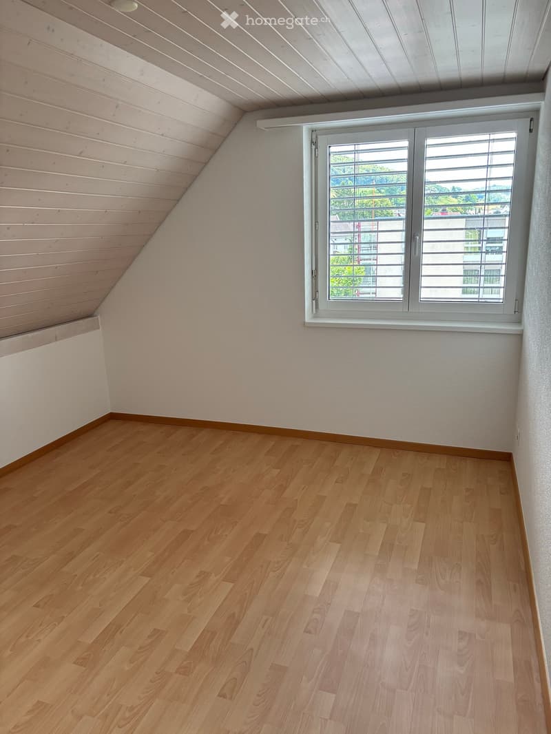 Empty room with wooden flooring, white walls, and a large window with blinds. The ceiling has exposed wooden beams.