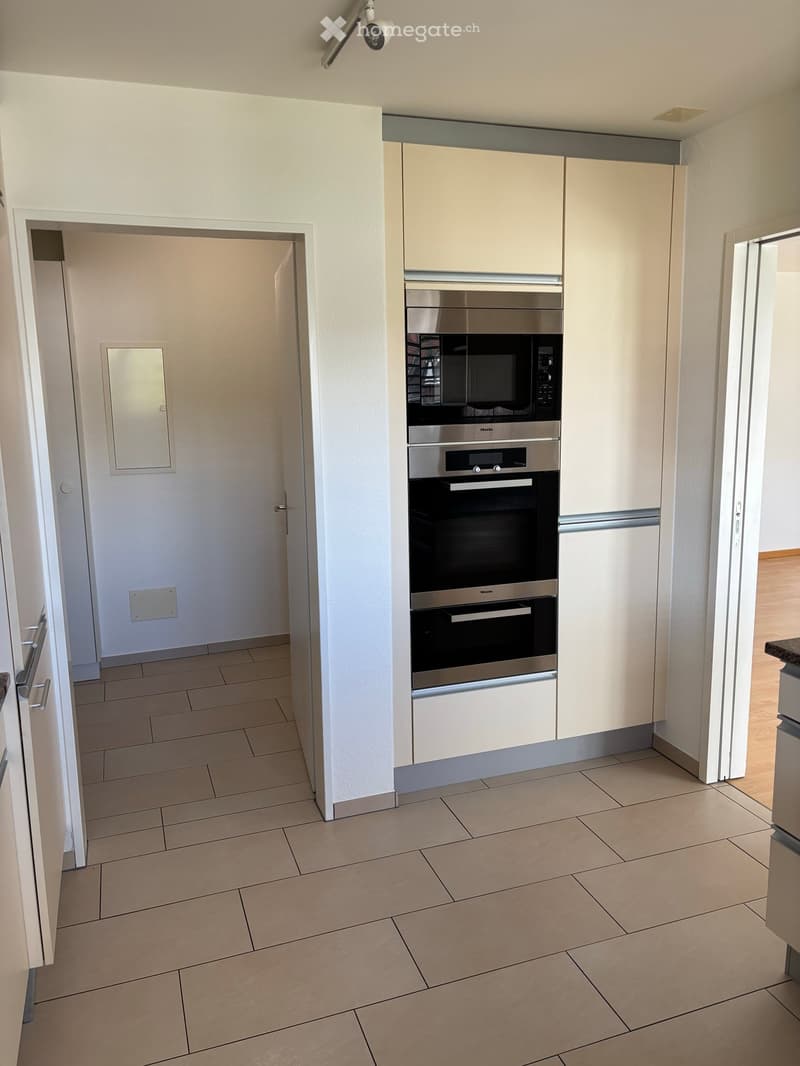 Modern kitchen with light-colored cabinets, stainless steel oven and microwave, tiled floor, and a doorway leading to another room.