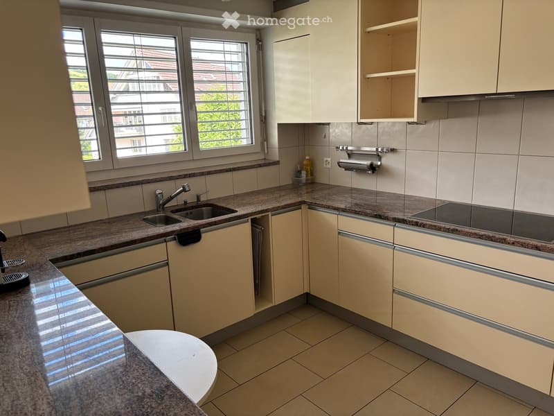 Modern kitchen with light-colored cabinets, granite countertops, and stainless steel appliances including a sink, stove, and dishwasher. The space is well-lit with large windows and has a tiled floor.