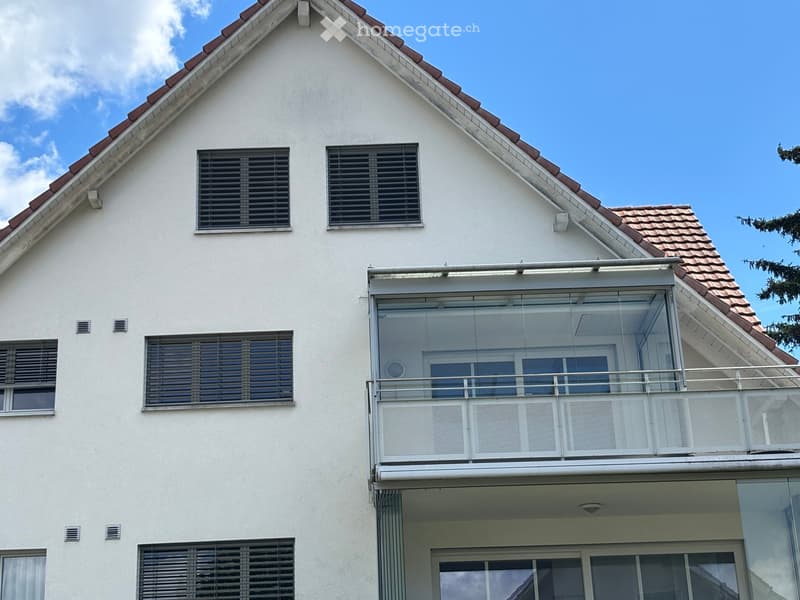 Two-story white building with a red-tiled roof, multiple windows with blinds, and a large balcony with glass railings.