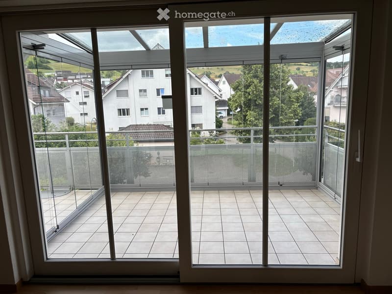 Balcony with tiled floor, glass railings, and a view of a residential area with houses and greenery.