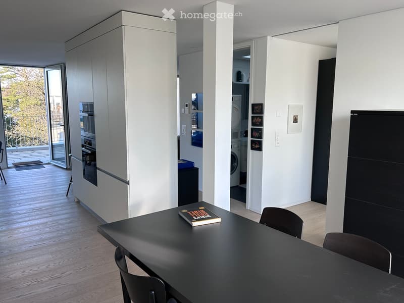 Modern, open-plan living and dining area with a black dining table, chairs, a cabinet, and a view to a balcony. A washing machine and fridge are visible in the adjacent space.