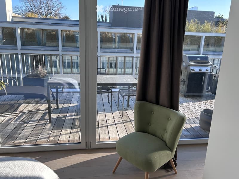 A modern, well-lit room with wooden flooring, a green upholstered chair, a small table, and a view of a balcony with a grill and outdoor furniture.