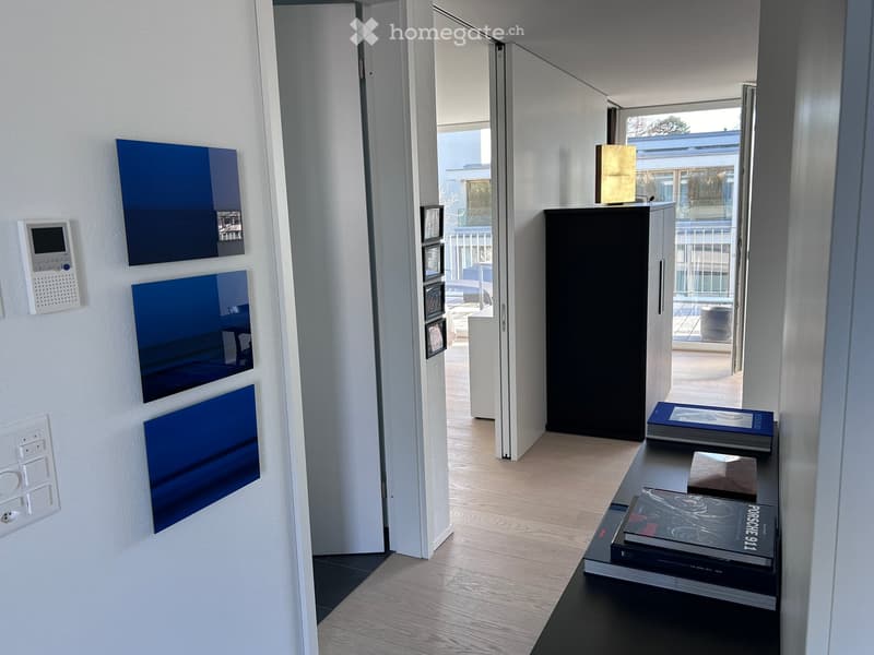 Modern hallway with light wooden flooring, white walls, and blue decorative panels. A small table holds books and a lamp, with a view to an adjacent room and balcony.