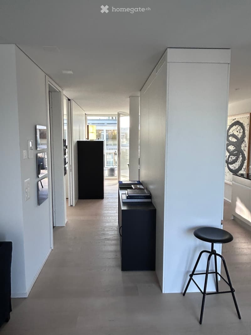 Modern hallway with light-colored walls, wooden flooring, and minimal furnishings including a stool and a small table with books.