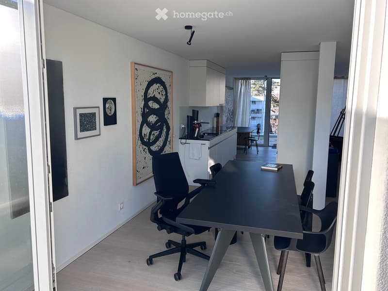 Modern, minimalist room with a black office chair, sleek table, laptop, and a small kitchen area with cabinets, sink, and a cup. Framed artwork adorns the walls.