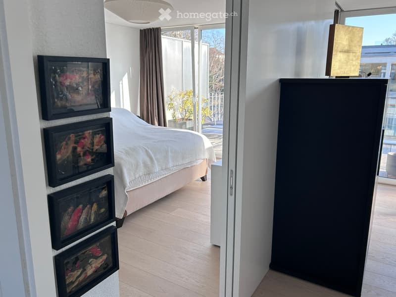 Modern bedroom with a large bed, framed artwork on the wall, and a tall wardrobe. The room features light wooden flooring and large windows with a view of the balcony.