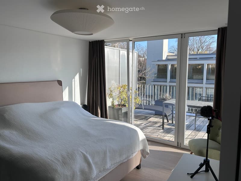 Bright bedroom with a large bed, a chair, a small table, and a balcony with outdoor furniture and potted plants.