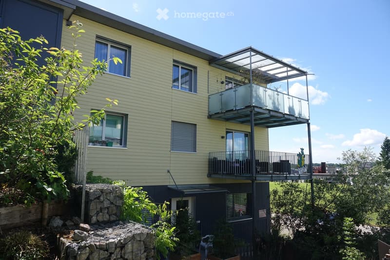 Modern two-story building with light yellow siding, multiple windows, and two balconies. The lower balcony has a glass enclosure, while the upper balcony is open with metal railings. The building is surrounded by greenery and a stone wall.