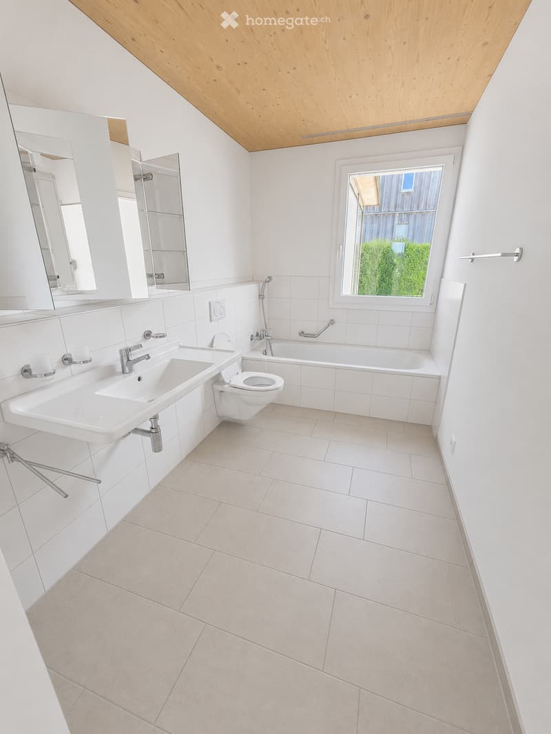 Modern bathroom with white tiles, a large sink, toilet, bathtub, and a mirrored cabinet. Features a skylight window and a wooden slanted ceiling.