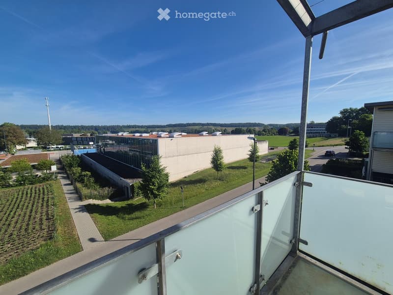 Balcony with glass railing overlooking a landscaped area with buildings, trees, and a clear blue sky.