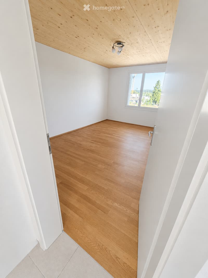 Empty room with wooden flooring, white walls, and a slanted wooden ceiling. Features a large window with a view of trees and a cityscape.