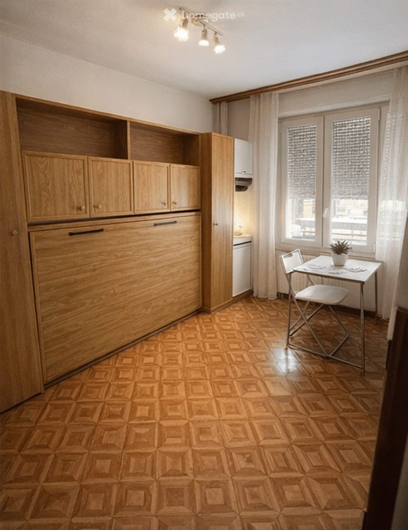 A compact, well-furnished room with wooden cabinets, a small table and chair, a potted plant, and a refrigerator. The room features wooden flooring and large windows with curtains.