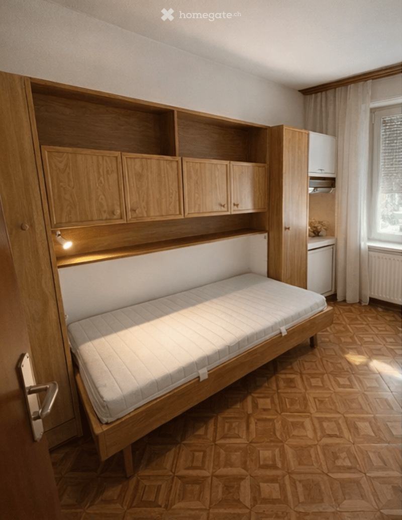 A compact bedroom with a single bed, built-in wooden wardrobe, and small cabinet. The room features wooden flooring and large windows with curtains.