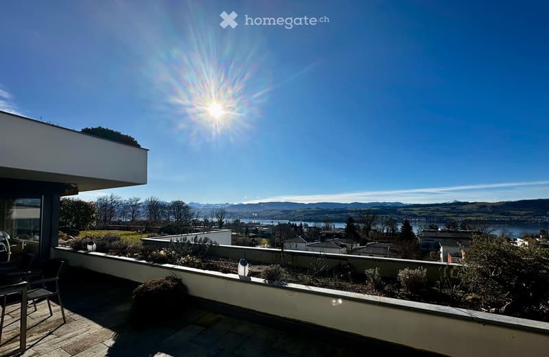 Balcony with a scenic view of a lake and mountains, featuring a chair and table, surrounded by low plants and shrubs.