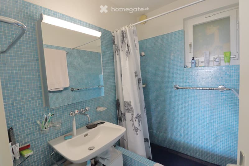 A small bathroom with blue tiled walls and floor, featuring a white sink with a chrome faucet, a mirror with a built-in light, a towel on a rack, a shower curtain, and various toiletries on a shelf.