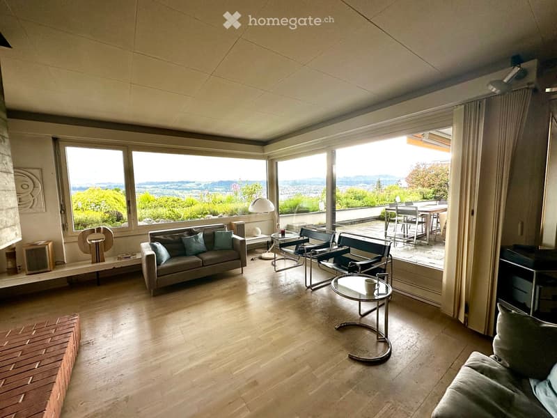 Modern living room with large windows offering scenic views, featuring a grey sofa, coffee table, chairs, and a lamp.