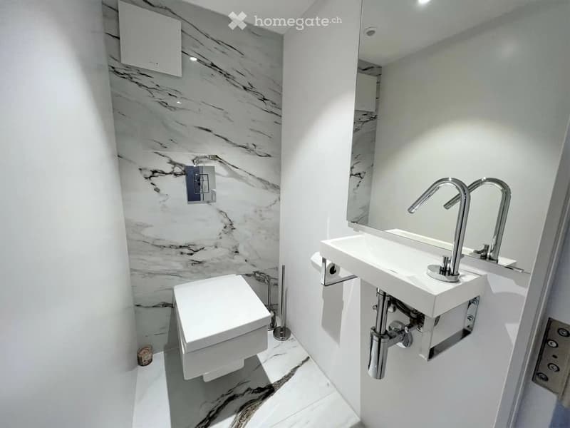 Modern bathroom with white marble walls and floors, featuring a toilet, a sink with a modern faucet, and a large mirror.