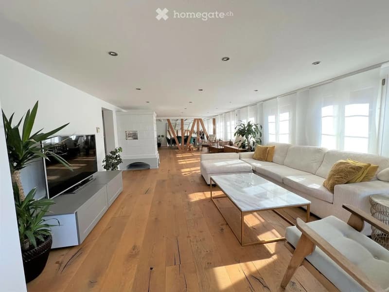 Bright living room with white walls, wooden flooring, large windows, a white sofa, coffee table, armchair, fireplace, TV stand, and potted plants.