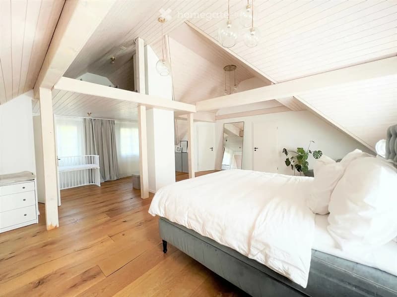 Bright, airy bedroom with wooden floors, white walls, and exposed wooden beams. Features a large bed with white bedding, a nightstand, a mirror, a small plant, and a baby crib in the background.