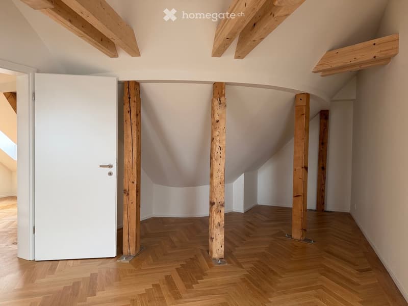 Empty hallway with wooden herringbone flooring, exposed wooden beams, and white walls. A white door is visible on the left side.