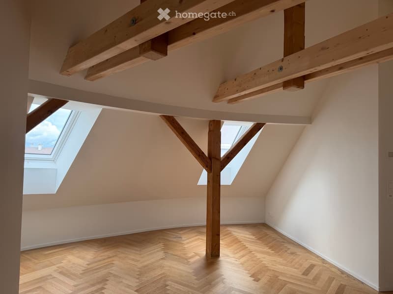 Empty attic room with wooden herringbone flooring, exposed wooden beams, and skylight windows allowing natural light.