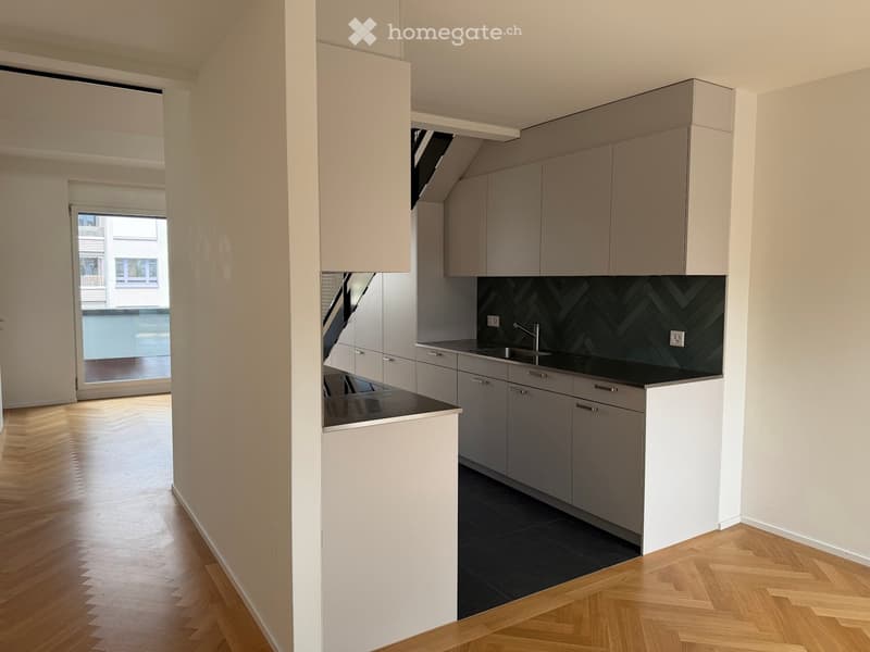 Modern kitchen with grey cabinets, black countertop, and a sink. The floor is wooden, and there is a view through a large window.