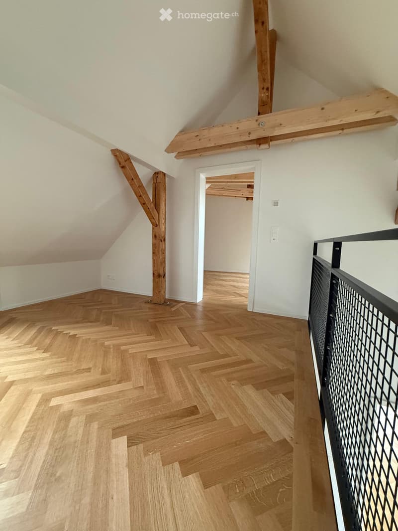 Spacious hallway with wooden herringbone flooring, white walls, exposed wooden beams, and a modern black metal railing leading to another room.