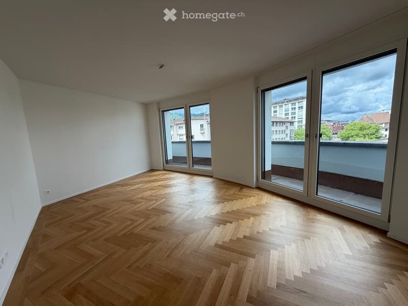 Empty room with wooden herringbone flooring, large windows, and white walls.