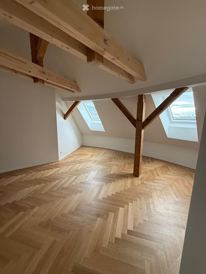 Empty attic space with wooden herringbone flooring, exposed wooden beams, and skylight windows allowing natural light.