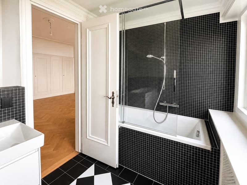 Modern bathroom with black tiled walls and floor, white bathtub with glass shower enclosure, white sink, and wooden flooring visible in adjacent room.